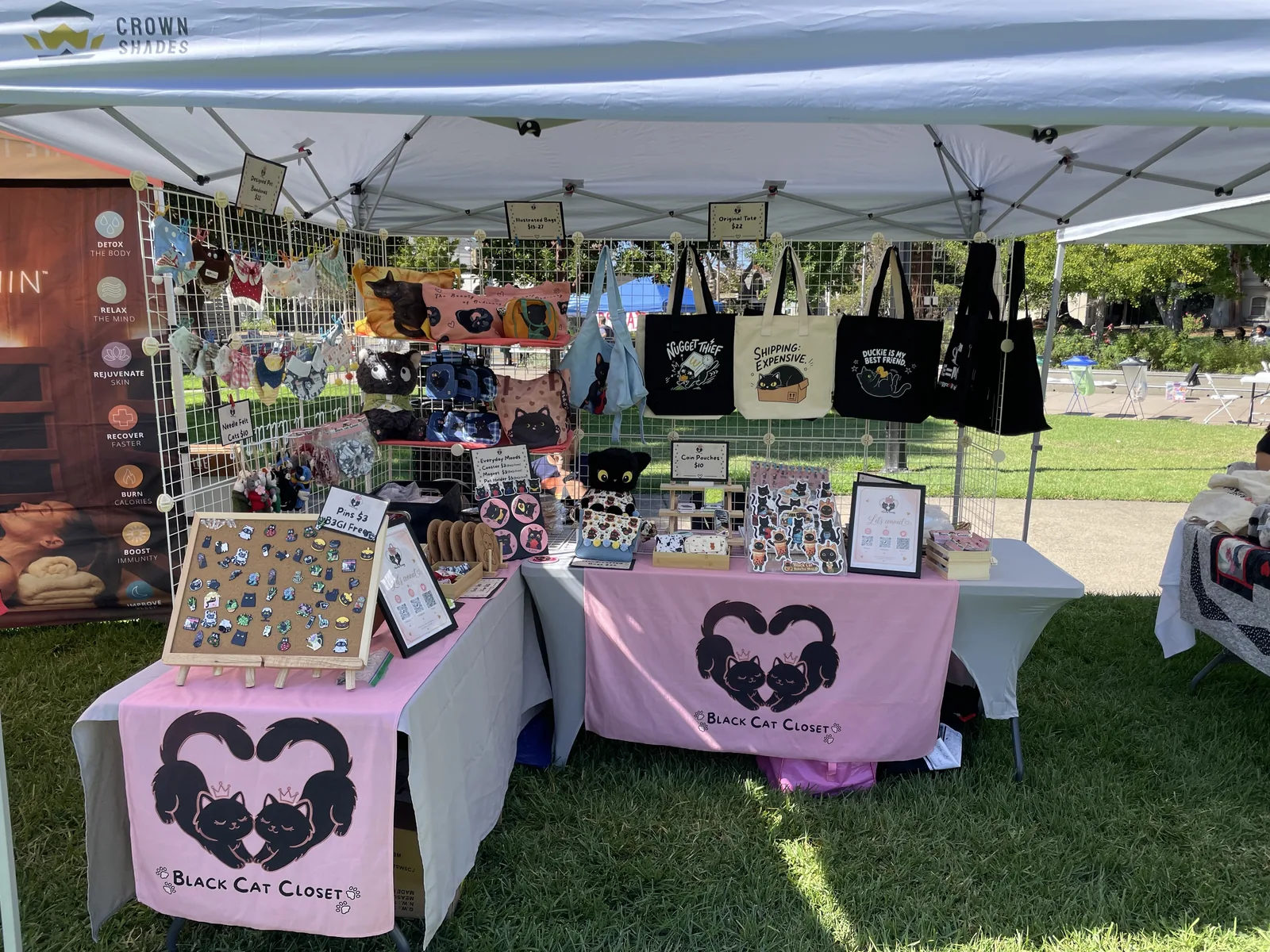 The Nyangi Studio USA booth on the lawn at Bizerkeley Food Fest, with cat tote bags on the grid wall, an enamel pin board, and pink table banners