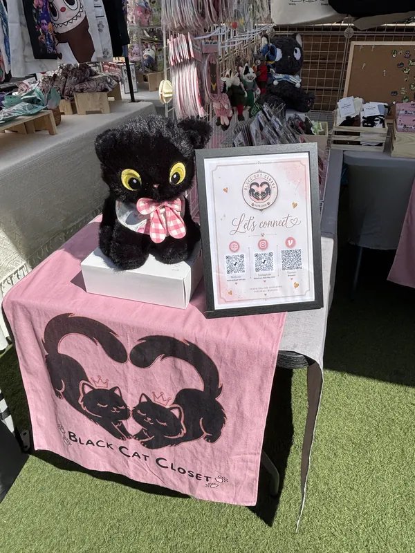 A black cat plush in a pink gingham bow beside our "Let's connect" QR sign