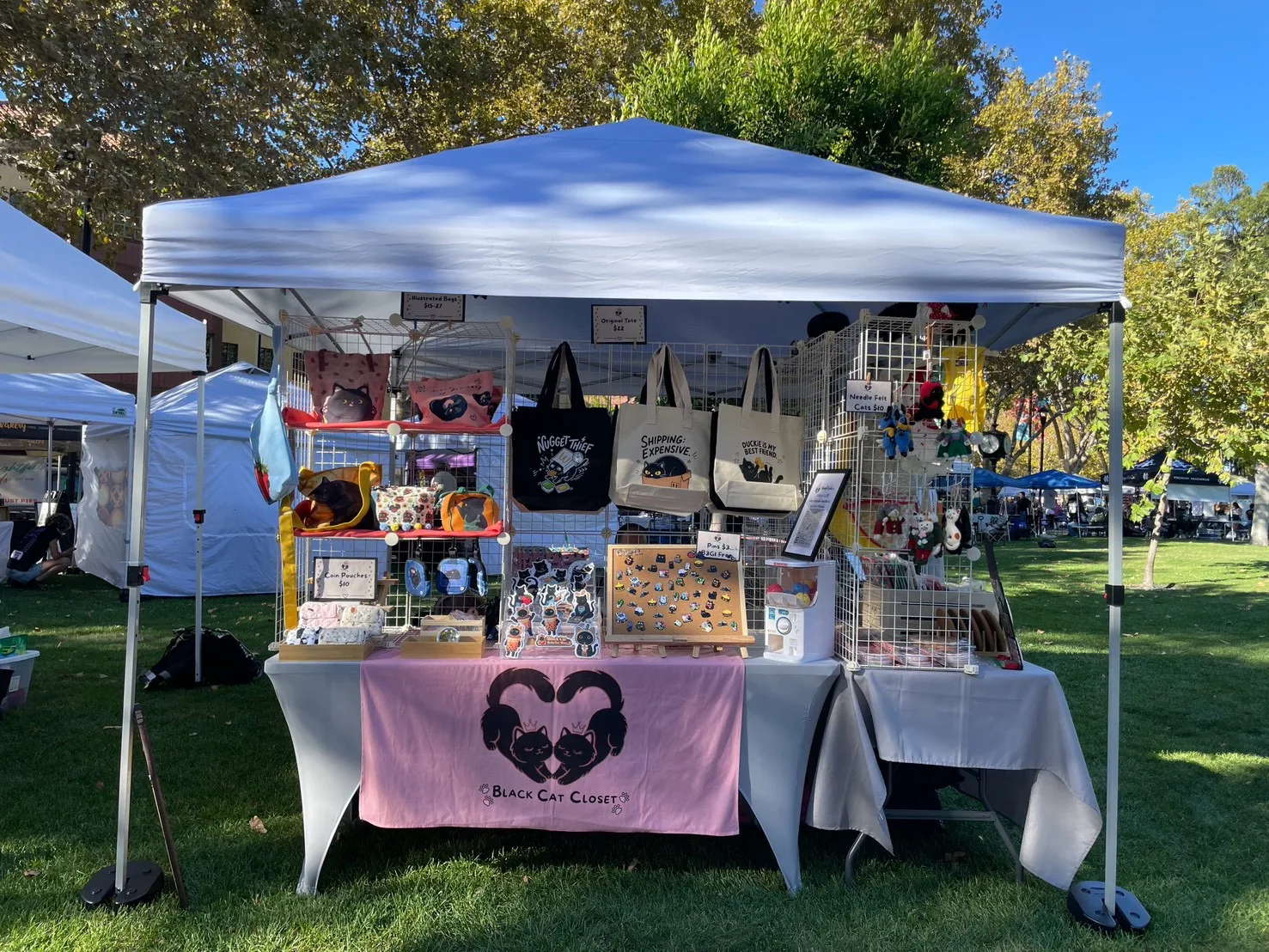 The Nyangi Studio USA booth under a white tent on the lawn at Todos Santos Plaza, with cat tote bags on the grid wall, an enamel pin board, and a pink table banner