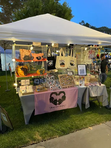 The booth after sunset, lit with warm string lights, showing illustrated bags on the shelves and the enamel pin board on its easel