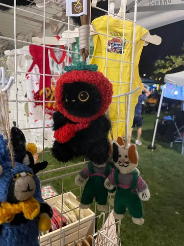 Wool-felt cat charms hanging on the grid wall at night, including a black cat in a red strawberry hood and a pair of calico cats in green overalls