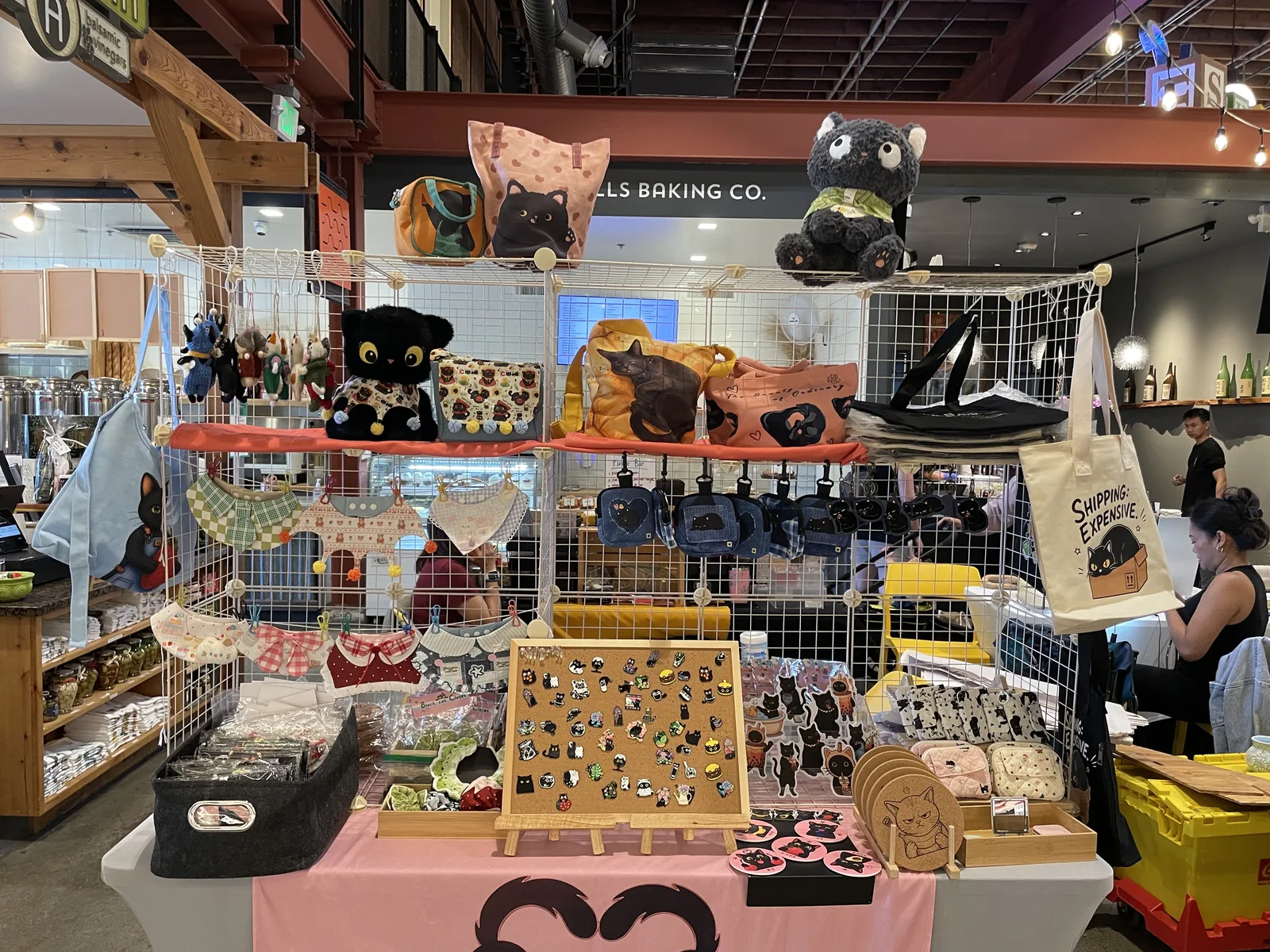 The Nyangi Studio USA booth at First Fridays, with cat totes, bandanas, denim pouches, and an enamel pin board on the marketplace floor