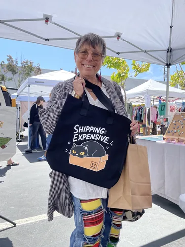 A happy customer holding up her new Shipping: Expensive cat tote bag at the booth
