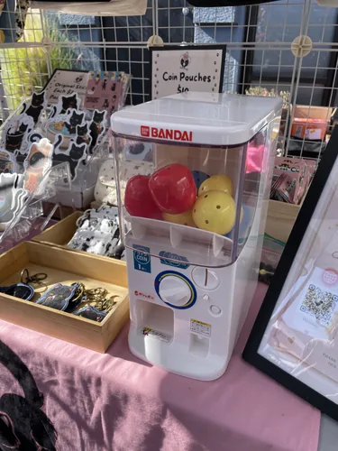 The Bandai gachapon capsule machine on the booth table next to keychains and the QR code sign