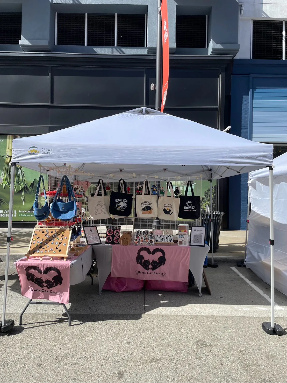 The Nyangi Studio USA booth under a white tent on the Bay Street promenade, with tote bags on the grid wall, an enamel pin board, and pink table banners