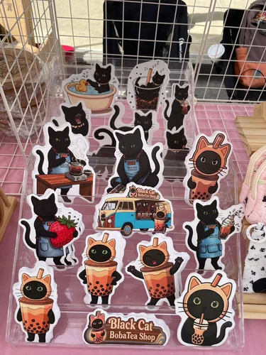 A clear acrylic stand full of Black Cat Boba Tea sticker sheets, with black cats in boba cup costumes, a cat boba-maker in a denim apron, and a boba van