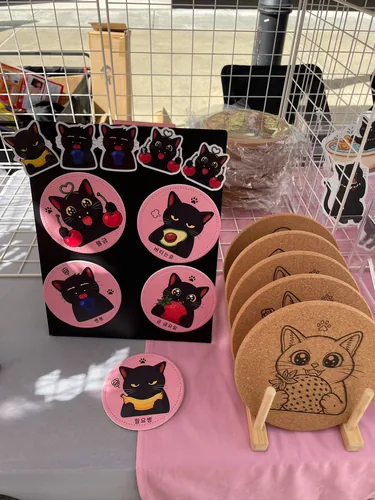 Pink Korean-text black cat coasters on a black display board with matching stickers, beside a stack of cork cat coasters on a wooden stand
