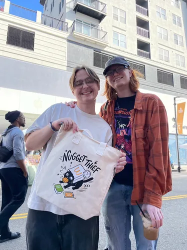 Two friends smiling, one holding up a natural canvas Nugget Thief tote