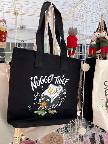 The Nugget Thief tote in black, hanging on the grid wall beside wool-felt cat charms in red and green outfits