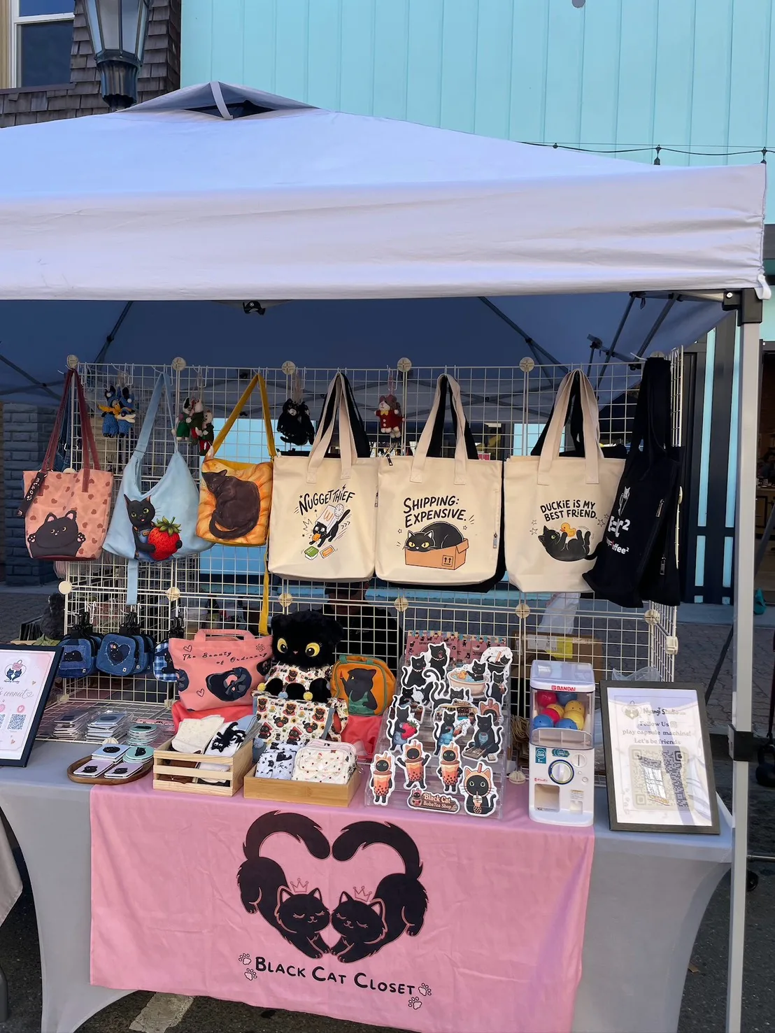 The Nyangi Studio USA booth under a white tent on Main Street in Martinez, with tote bags and shoulder bags on the grid wall, a capsule machine, and a pink table banner