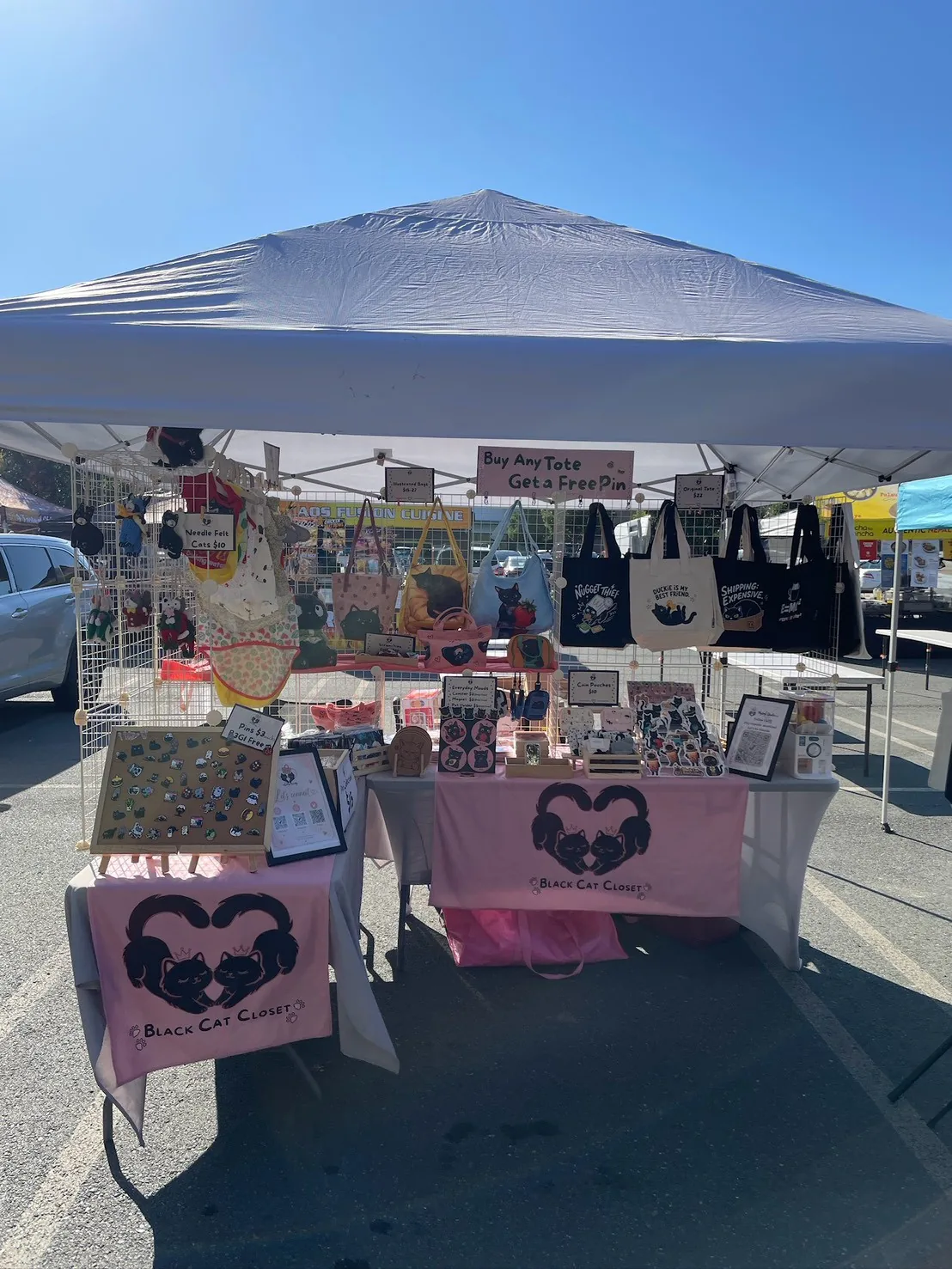 The Nyangi Studio USA booth under a white tent on a sunny afternoon, with illustrated bags and cat tote bags on the grid wall, an enamel pin board on an easel, and pink table banners
