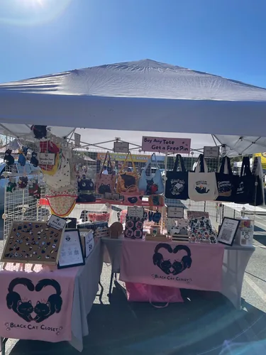 The full booth in bright late-afternoon sun, with the Buy Any Tote Get a Free Pin sign above the bag wall and wool-felt cats hanging on the left