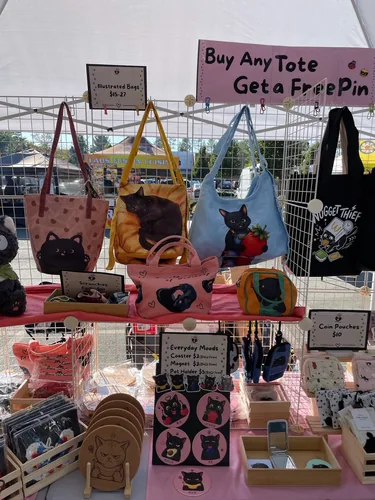 Illustrated black cat bags on the grid wall — a pink heart tote, a sleeping cat on a yellow bag, and a blue shoulder bag with a cat hugging a strawberry — above Everyday Moods coasters and magnets