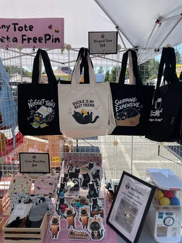 Our original tote bags in a row — Nugget Thief, Duckie Is My Best Friend, Shipping: Expensive, and E=MC² — above coin pouches, Black Cat Boba Tea stickers, and the capsule machine